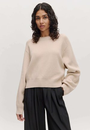 Beiger Pullover mit Rundhalsausschnitt, gerippten Bündchen und Saum. Weicher, leicht strukturierten Stoff. Kombiniert mit lockeren, schwarzen, plissierten Hosen.