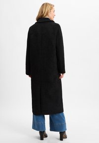 Langer schwarzer Mantel aus strukturiertem Stoff, gerader Schnitt und breite Revers. Getragen über ausgestellten blauen Jeans und schwarzen Stiefeln mit Absatz.