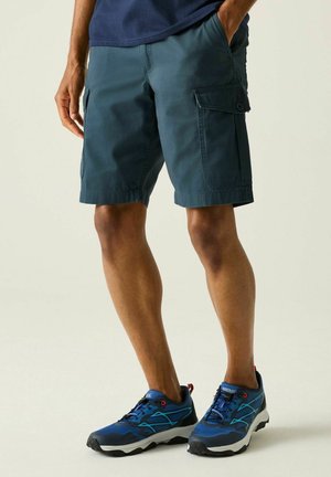 Man die donkergroene cargoshorts en blauwe sportschoenen draagt, staat met één hand in de zak tegen een lichte achtergrond.