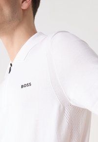 Polo blanc avec col, arborant un logo "BOSS" noir. Panneau en maille texturée sur l'épaule pour une meilleure respirabilité. Encolure zippée.