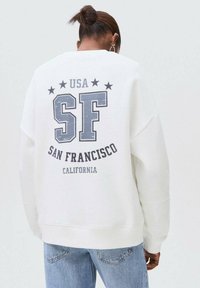 Oseba z rjavimi lasmi, zvezanimi v sok, nosi belo majico s tiskom "USA SF San Francisco California" na hrbtni strani in svetlo modre kavbojke.