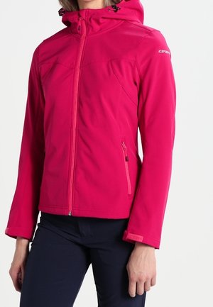 Frau trägt eine leuchtend pinke Kapuzenjacke mit seitlichen Reißverschlusstaschen und marineblaue Hose vor einfarbigem Hintergrund.