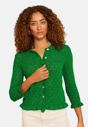 Woman with curly hair wearing a green textured cardigan with gold buttons and cream pants, holding the cardigan near the chest.