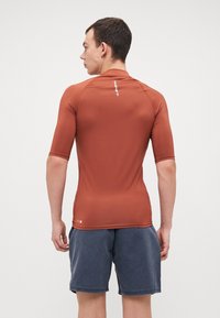 Brun, kortermet rash guard med elastisk stoff, glatt tekstur og flate sømmer, brukt med blå shorts. Logo synlig på ryggen.
