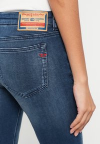 Gros plan sur la poche arrière bleue d’un jean Diesel et l’écusson du logo sur la ceinture, la main droite de la personne reposant le long du corps.