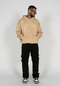 Beige oversized hoodie med grafisk print, sorte cargo-bukser og sort-hvide sneakers. Model iført solbriller, står imod en neutral baggrund.