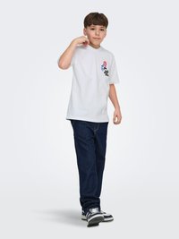 Jongen in een wit T-shirt met print, donkerblauwe spijkerbroek en sneakers, staand met één hand bij zijn nek tegen een effen lichte achtergrond.