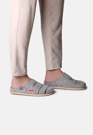 Pantofole in lana grigia con design a strisce e ricami rossi, dotate di un retro aperto e suola simile al giunco. Indossate con pantaloni beige.