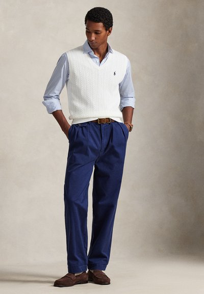 Λευκό πλεκτό γιλέκο πάνω από μπλε και λευκό πουκάμισο με καρό, συνδυασμένο με μπλε σκούρο πλισέ παντελόνι και καφέ σουέτ παπούτσια. Ουδέτερο φόντο.