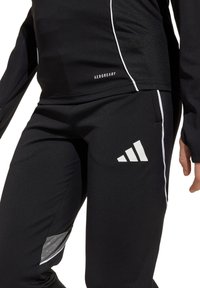 Schwarze Sportbekleidung aus feuchtigkeitstransportierendem Material, mit schlankem Design, weißen Akzenten und einem Adidas-Logo. Die Hose verfügt über reflektierende Details.