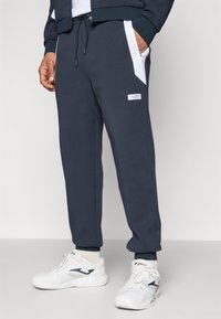 Marine joggebukser laget av myk stoff, med hvite sidestykker, snor i midjen og et frontlogo merke. Kombinert med hvite joggesko.