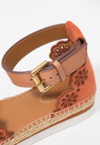 Sandal in suede arancione con cinturino alla caviglia in pelle beige, fibbia dorata, intagli floreali, dettaglio in juta intrecciata e suola in gomma bianca.