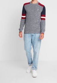 Pull en maille bleu marine et gris avec des accents rayés rouges et blancs sur les manches, un col rond et des poignets côtelés, associé à un jean bleu clair.
