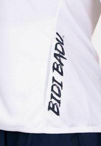 Tissu blanc avec un texte vertical noir indiquant "BIOT BADU", partiellement visible le long d'une couture au-dessus de vêtements foncés.