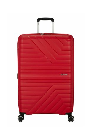 Valise rigide rouge à rainures diagonales, quatre roues pivotantes, poignée noire extensible, et logo "American Tourister" sur le devant.