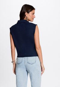 Femme portant un haut sans manches coloris navy avec col et un jean taille haute bleu clair, de dos avec le visage tourné sur le côté, sur fond blanc.