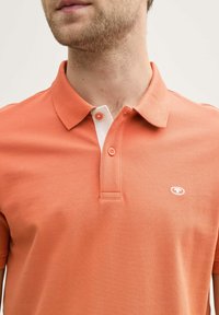 Orange polo tröja med texturerad tyg, med en vit krage och en enda knäppning vid halsen, logotyp på vänster bröst.