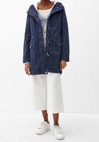 Parka azul marino con capucha forrada de piel, dos bolsillos frontales y cordones ajustables. Combinada con pantalones de pierna ancha blancos y zapatillas.