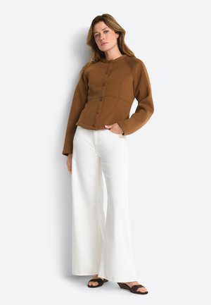 Kvinde iført en brun jakke med knapper og lange ærmer, hvide vidde bukser og sorte åbne hæle, der står mod en hvid baggrund.