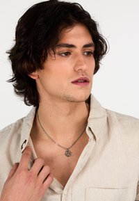 Collana in argento con un pendant geometrico indossata su una camicia di lino beige chiaro, con colletto rilassato e dettaglio dei bottoni.