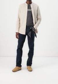 Beige linen button-up shirt over a gray t-shirt, dark blue denim jeans, and tan suede sneakers. Black bandana tied at waist.