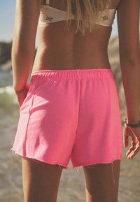 Rosa bomullsshorts med en rå fåll och resårmidja, visade bakifrån mot en strandbakgrund.