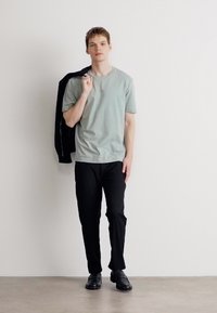 Light green t-shirt, slim-fit black pants, and black shoes. The model holds a black jacket over his shoulder against a neutral background.