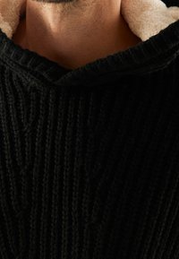 Svart, stickad, huva tröja med ett strukturerat mönster och en krämfärgad fleecefoder vid halsringningen. Närbild av tyget.