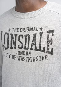 Világosszürke kötött pulóver, kerek nyakkivágással. Fekete szöveges grafika olvasható rajta: "Lonsdale London Westminster város" elhasználódott stílusban.