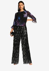 Blusa nera trasparente con motivo galattico, scollatura plissettata; pantaloni a gamba larga neri con paillettes e frange lunghe, abbinati a una piccola borsa nera a tracolla.