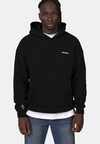 Sweatshirt à capuche noire en mélange de coton, avec une poche avant, un logo sur la poitrine et des poignets côtelés. Porté par-dessus un t-shirt blanc.
