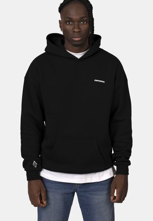 Black hooded sweatshirt made of cotton blend, featuring a front pocket, a logo on the chest, and ribbed cuffs. Worn over a white T-shirt.
