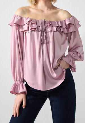 Femme portant un chemisier rose à épaules dénudées avec volants et manches longues, associé à un pantalon bleu foncé, main dans la poche, fond neutre.