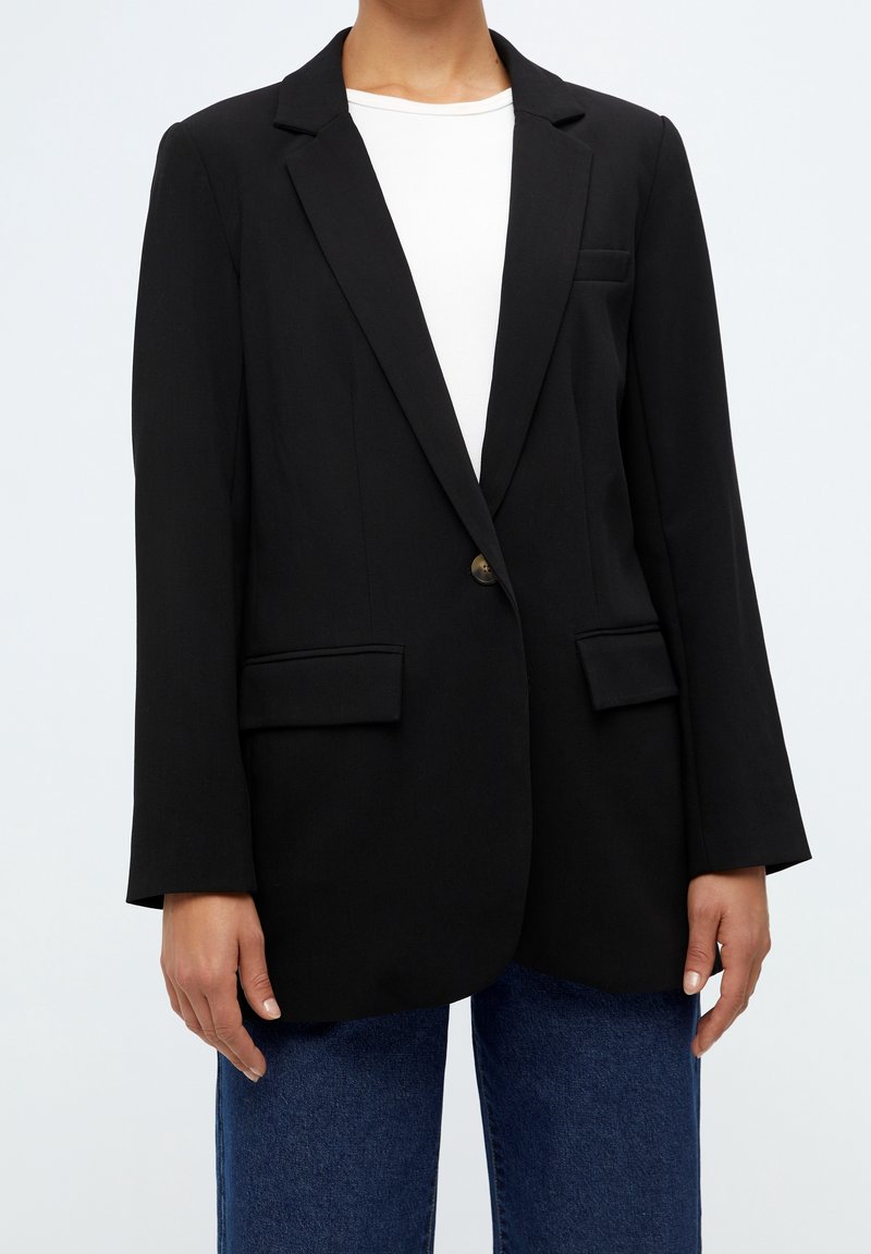 Personne portant un blazer noir à un bouton sur une chemise blanche avec un jean bleu, debout devant un fond clair uni.