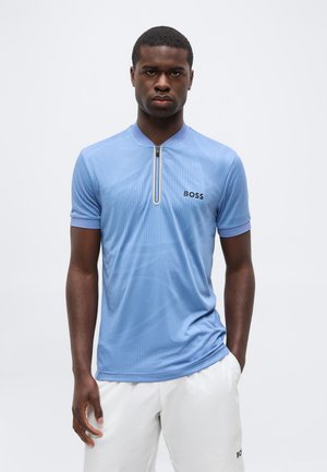 Uomo che indossa una camicia azzurro chiaro a maniche corte con zip e pantaloncini bianchi con loghi "BOSS", in piedi davanti a uno sfondo chiaro e uniforme.