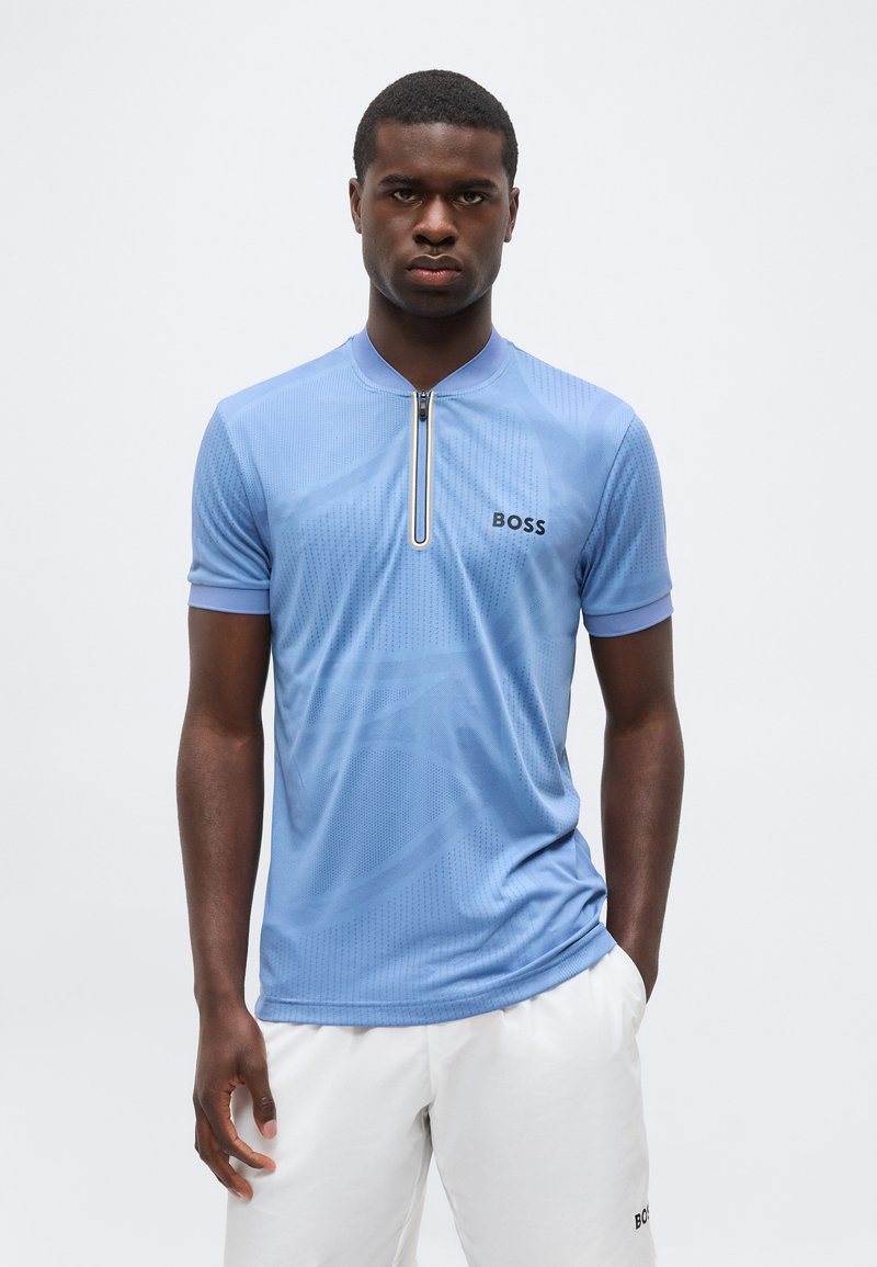 Άνδρας με ανοιχτό μπλε κοντομάνικο πουκάμισο με φερμουάρ και λευκά σορτς με λογότυπα "BOSS", στέκεται μπροστά σε απλό φωτεινό φόντο.