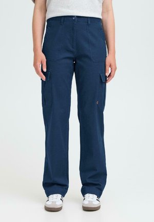 Person trägt navyblaue Cargo-Hosen mit Klappen-Taschen und weiße Sneakers und steht vor einem schlichten hellen Hintergrund.