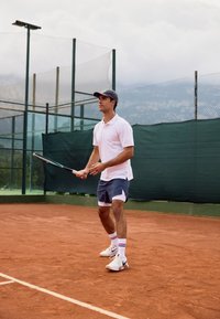 Un tennista in polo bianco e pantaloni corti grigi tiene una racchetta su un campo in terra battuta. Indossa un cappellino e scarpe da ginnastica bianche con strisce colorate.