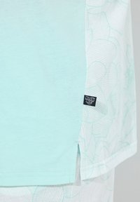 T-shirt verde menta con un motivo floreale sul lato, maniche corte e una piccola etichetta nera del marchio vicino all'orlo.