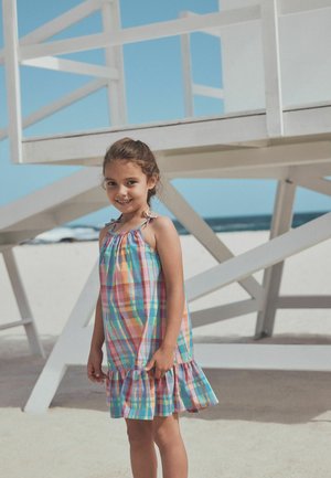 Smiling girl in a colorful plaid sundress standing near a white lifeguard tower on a sunny beach with ocean in the background.