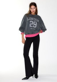 Felpa grigia oversize con stampa "LONDON 29", indossata sopra una maglia rosa a righe. Completa il look un paio di pantaloni neri svasati e décolleté nere con punta.