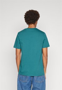 T-shirt corto in teal con collo a giro, realizzato in tessuto morbido, mostrato da dietro. Abbinato a jeans blu dal taglio morbido.