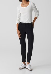 Jeans negros ajustados con tiro alto, combinados con una camiseta blanca de manga larga. Los jeans tienen trabillas para cinturón y bolsillos frontales; textura de tela suave.