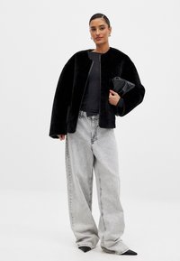 Svart konstläderjacka med rund halsringning, kombinerat med en åtsittande svart topp och stora ljusgrå denimjeans. Modellen håller en svart clutch.