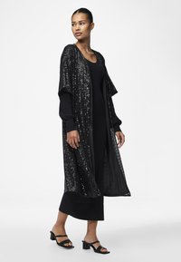 Kimono lung negru cu paiete, cu mâneci scurte, purtat peste o rochie neagră ajustată, asortat cu sandale cu toc bloc cu curea neagră.