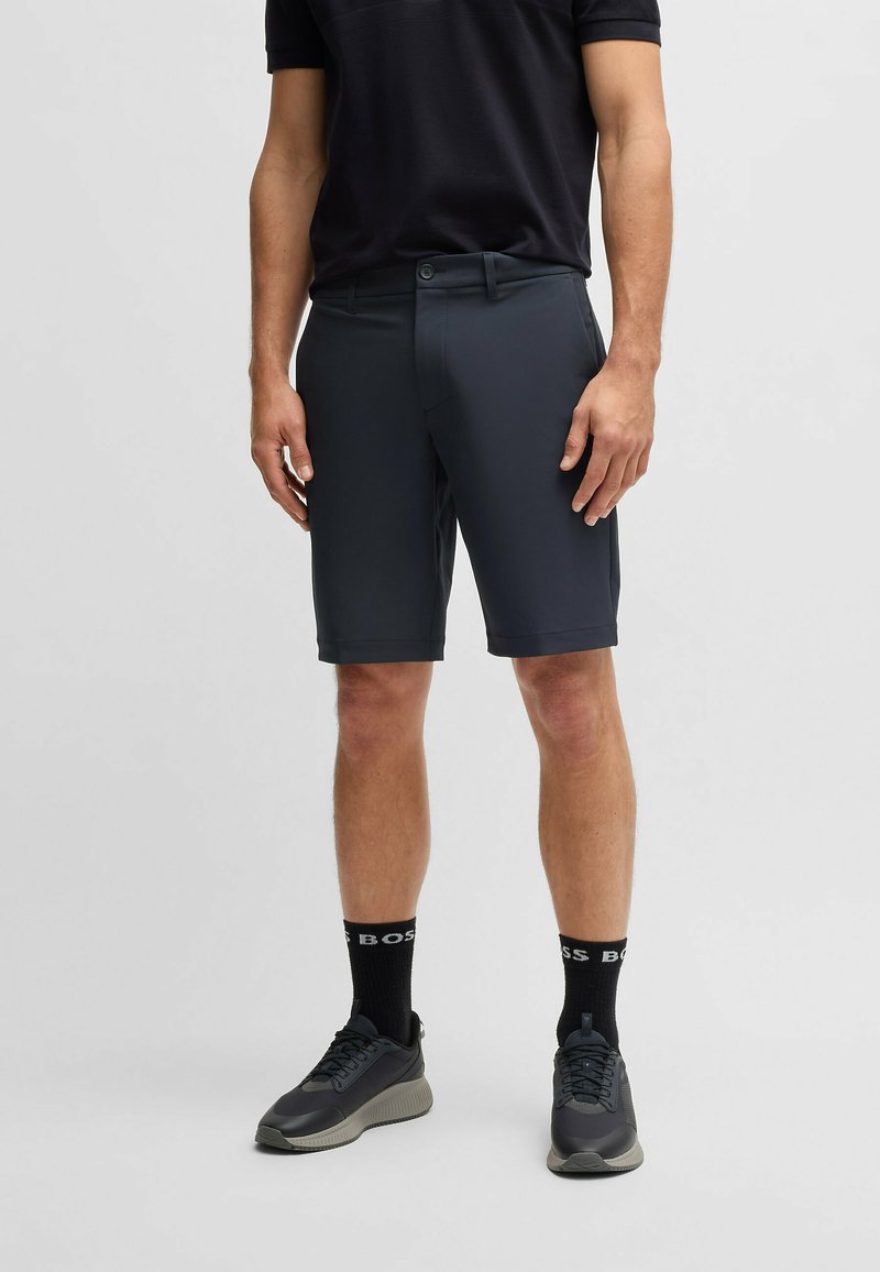 Pantaloni corti grigio scuro da uomo realizzati in un materiale liscio e leggero. Presentano una chiusura con bottone e tasche laterali, abbinati a sneaker nere e calze.
