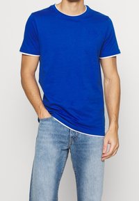 Niebieska bawełniana koszulka z białym wykończeniem przy kołnierzyku i rękawach. Posiada dyskretny, napisany pismem odręcznym logo na piersi oraz luźny krój.