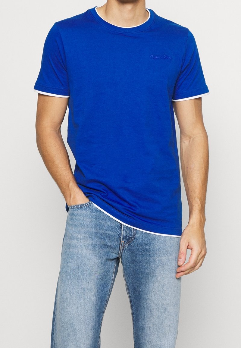 Niebieska bawełniana koszulka z białym wykończeniem przy kołnierzyku i rękawach. Posiada dyskretny, napisany pismem odręcznym logo na piersi oraz luźny krój.