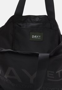 Czarna torba na ramię wykonana z tkaniny, z przestronnym wnętrzem, zapięciem na zatrzask oraz zieloną etykietą z nazwą marki i informacją o recyklingu.