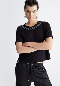 T-shirt nera corta con scollatura rotonda, caratterizzata da un motivo decorativo in argento attorno al collo. Tessuto liscio, maniche corte, design semplice.